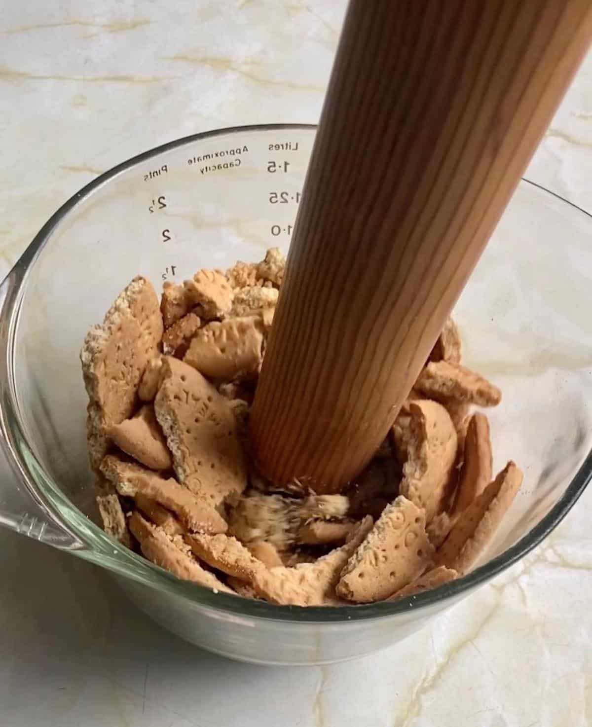 A bowl full of biscuits or cookies being crushed with the end of a rolling pin for the biscuit base