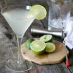 A martini glass with a pale green cocktail inside and a chopping board with limes on.