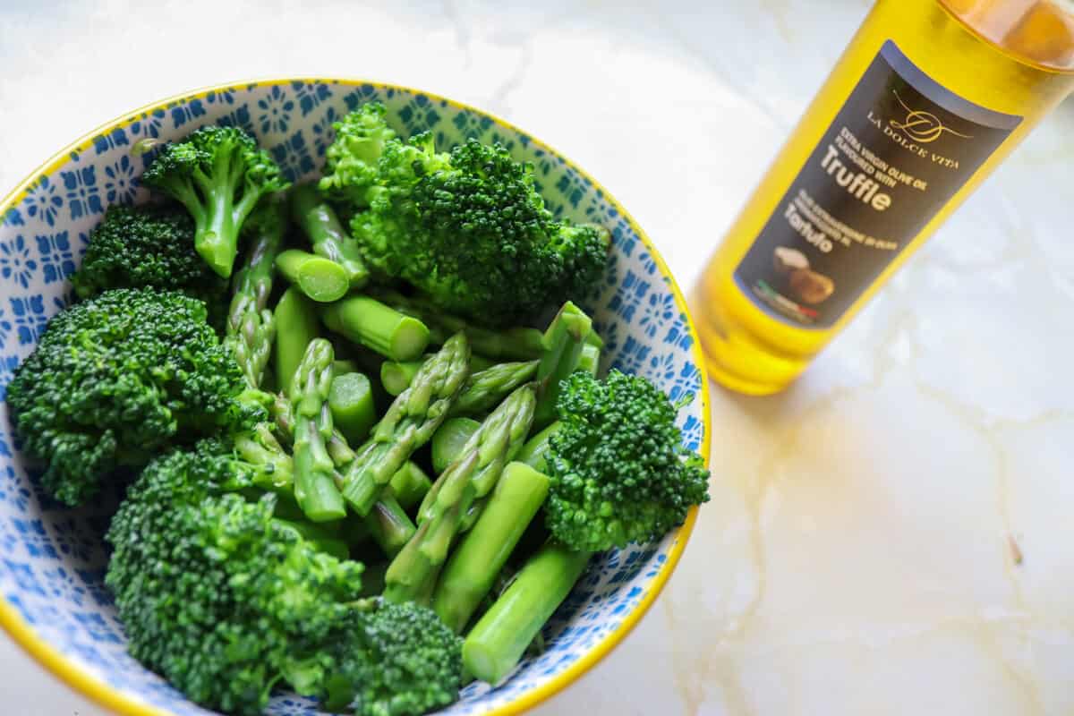 A bowl of cooked broccoli and asparagus and a bottle of truffle oil 