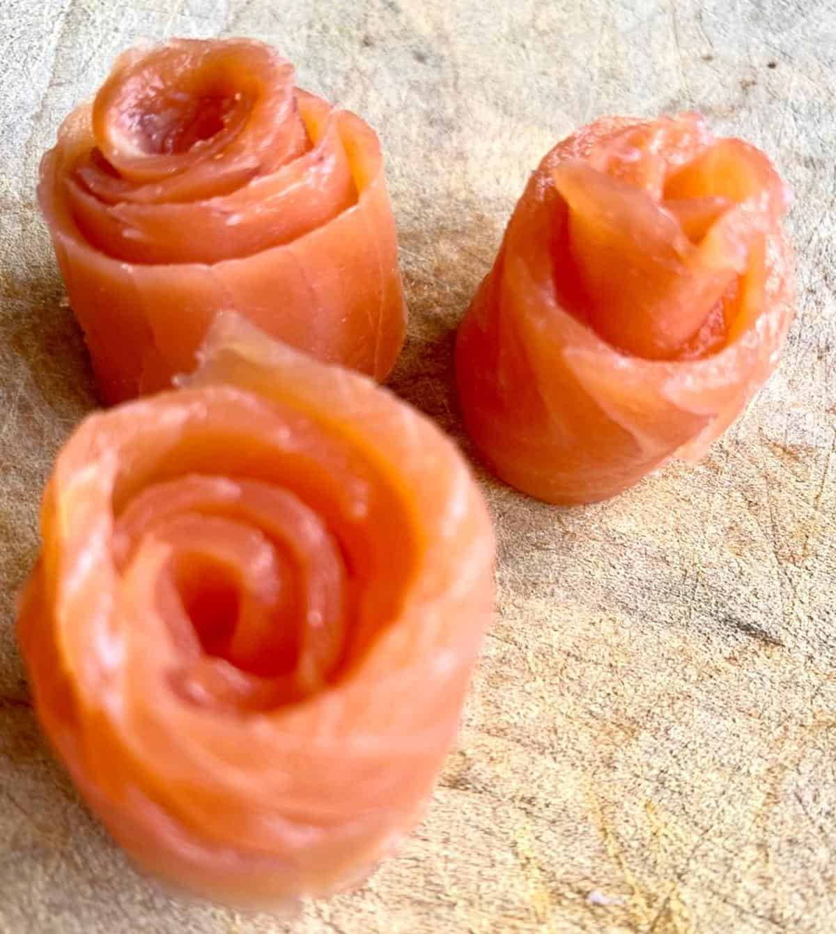 Pieces of smoked trout curled into rose shapes on a wooden chopping board. 