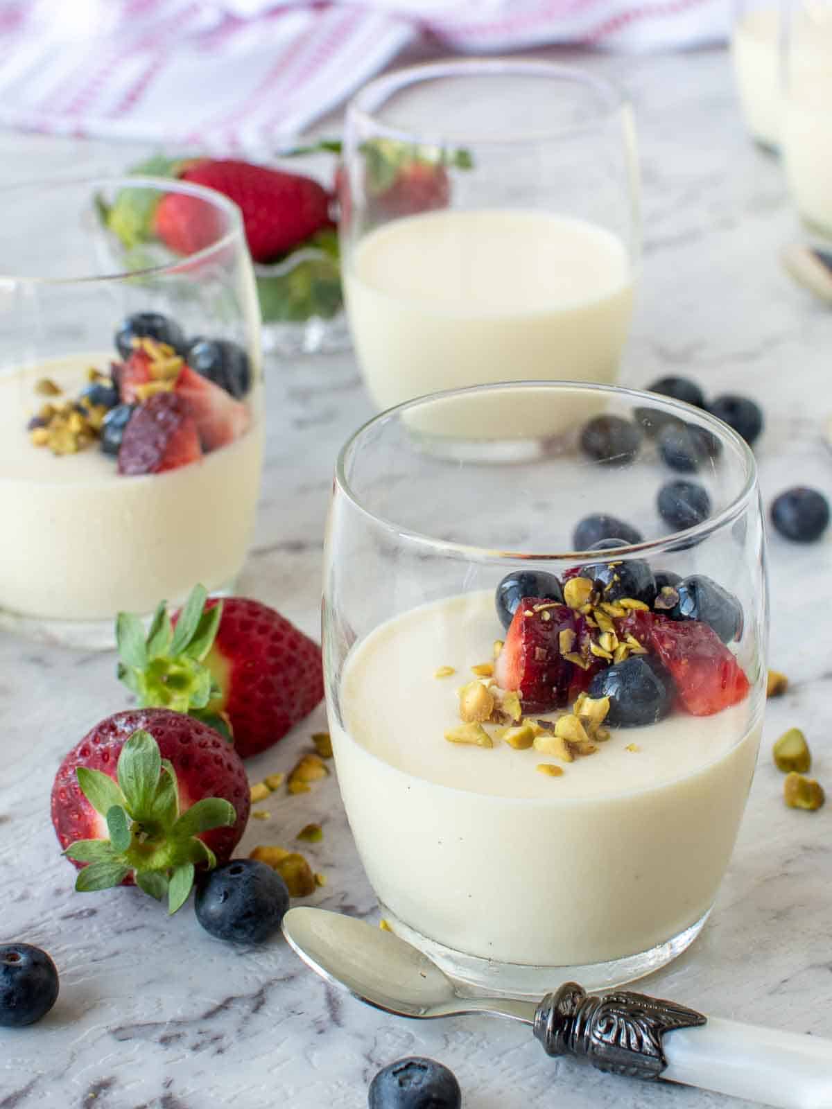 3 drinking glasses filled with panna cotta that is topped with fresh berries and pistachio nuts