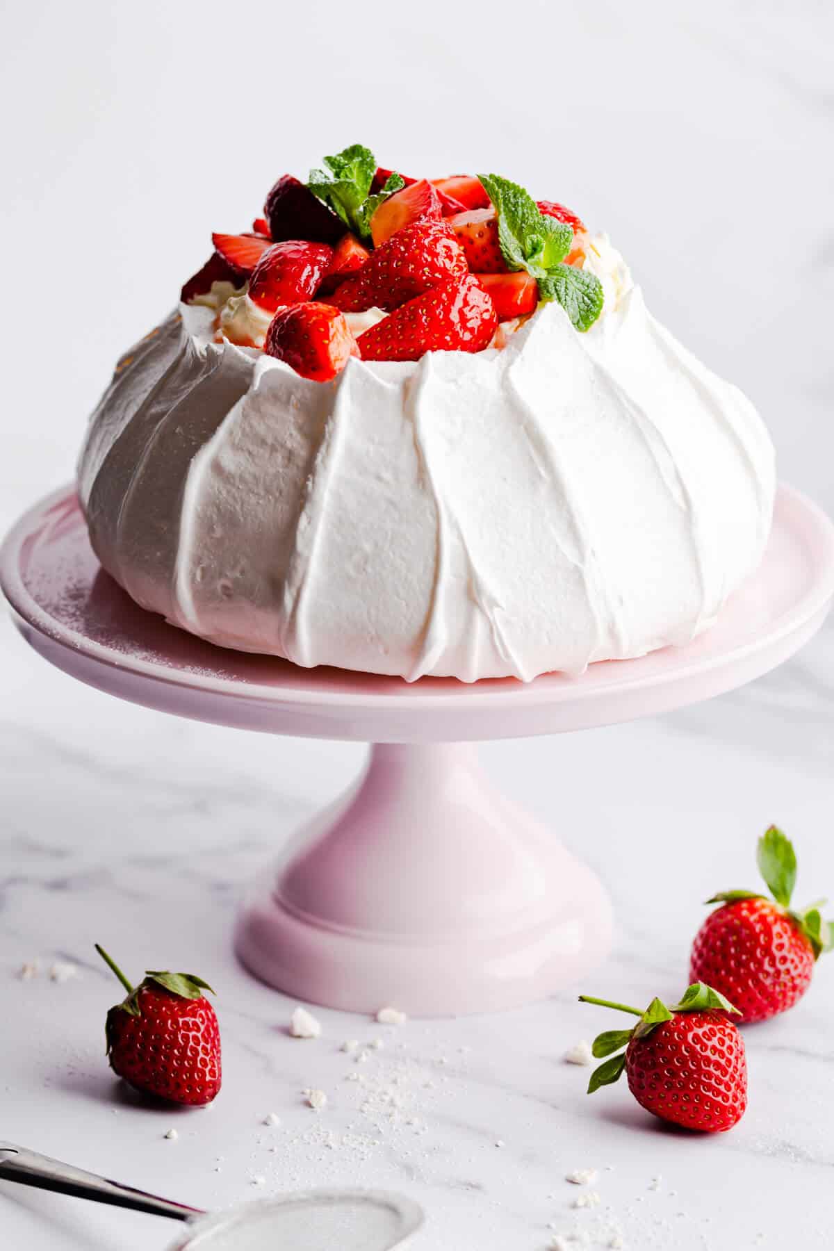 A cake stand with a strawberry pavlova on it, surrounded by fresh strawberries.
