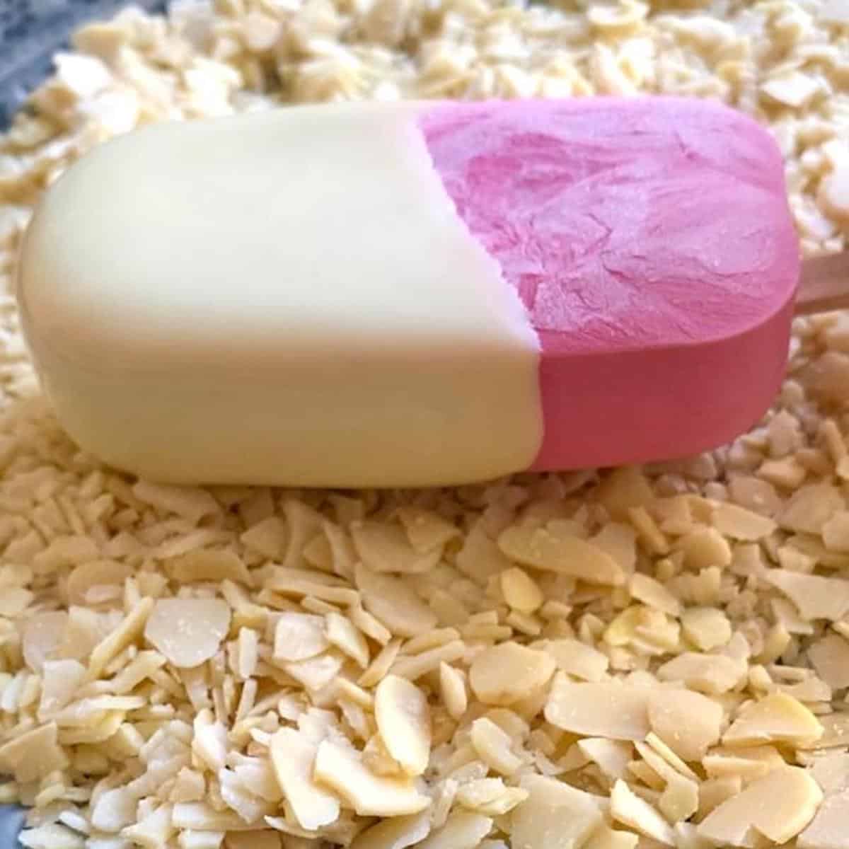 A raspberry ice cream bar coated in white chocolate being dipped into crushed flaked almonds on a plate.