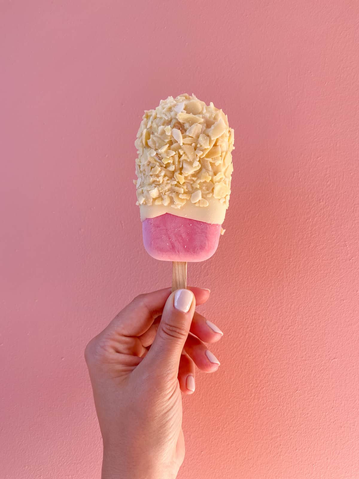 A hand holding up a pink raspberry ice cream bar that has been dipped in white chocolate and flaked almonds.