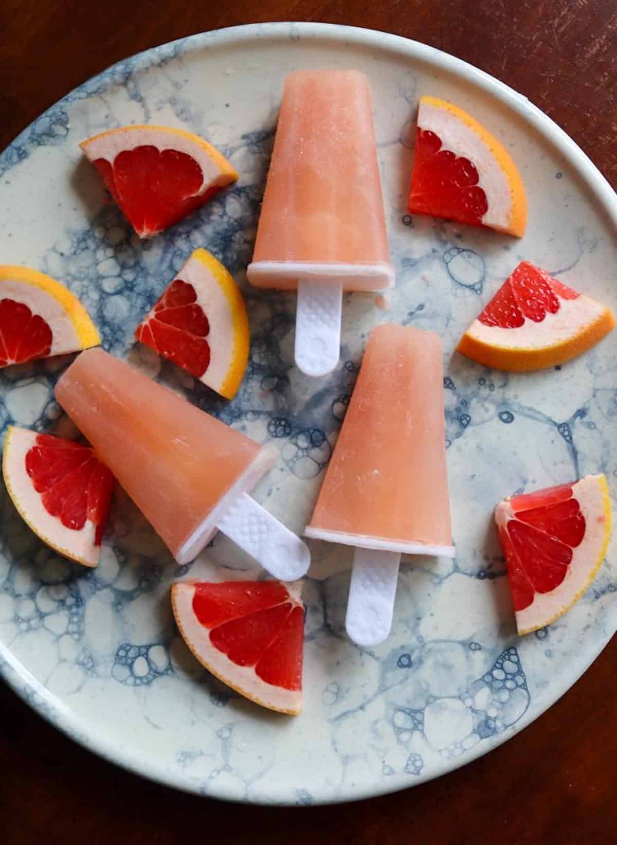 Vodka greyhound frozen cocktail popsicles on a platter surrounded by wedges of fresh grapefruit.