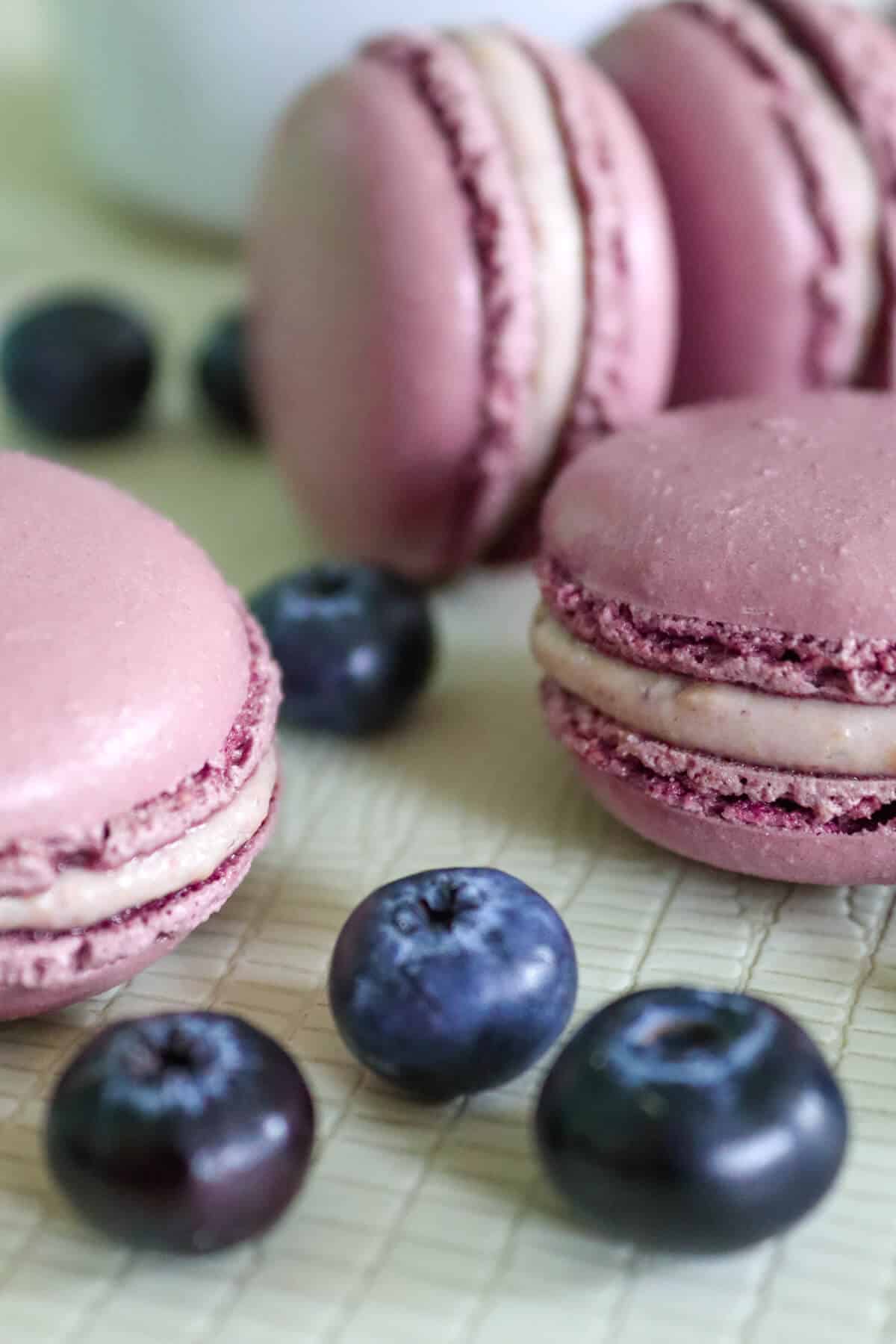 Blueberrry cheesecake macarons on a table with fresh blueberries. 