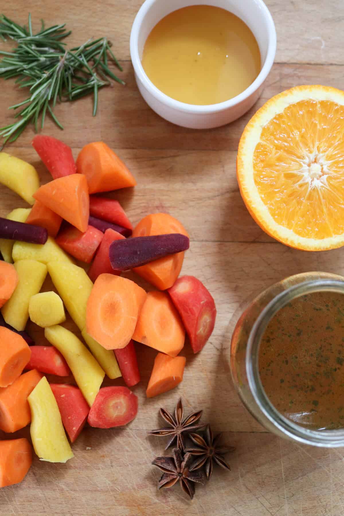 Ingredients on a chopping board: rainbow carrots, honey, vegetable stock, rosemary, star anise and orange. 