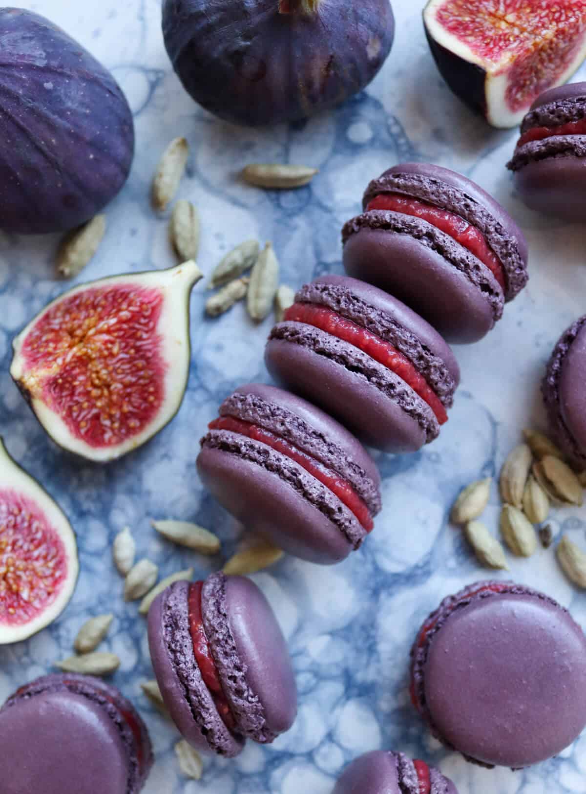 Fig and cardamom macarons on a platter surrounded by fresh figs and cardamom pods. 