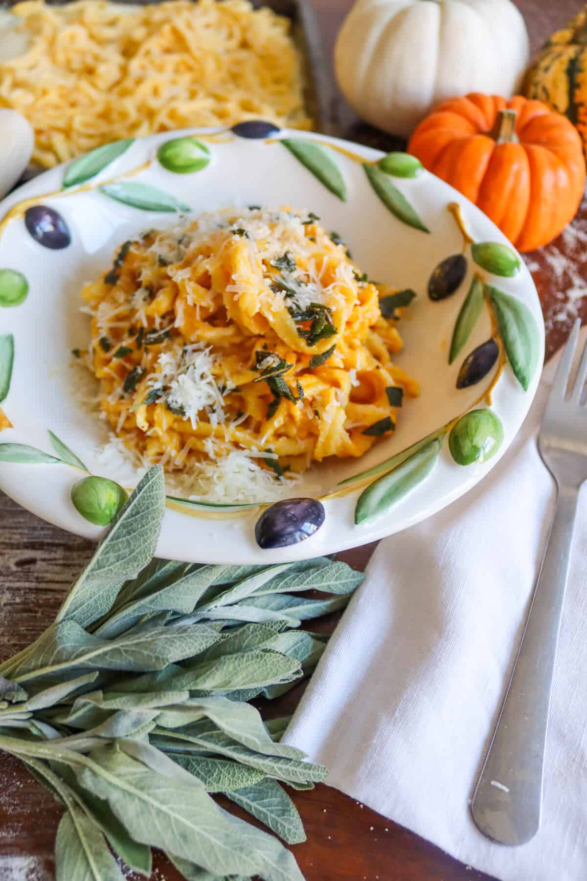 A bowl of creamy sage and pumpkin tagliatelle next to a tray of fresh pasta, a bunch of fresh sage and some ornamental pumpkins. 