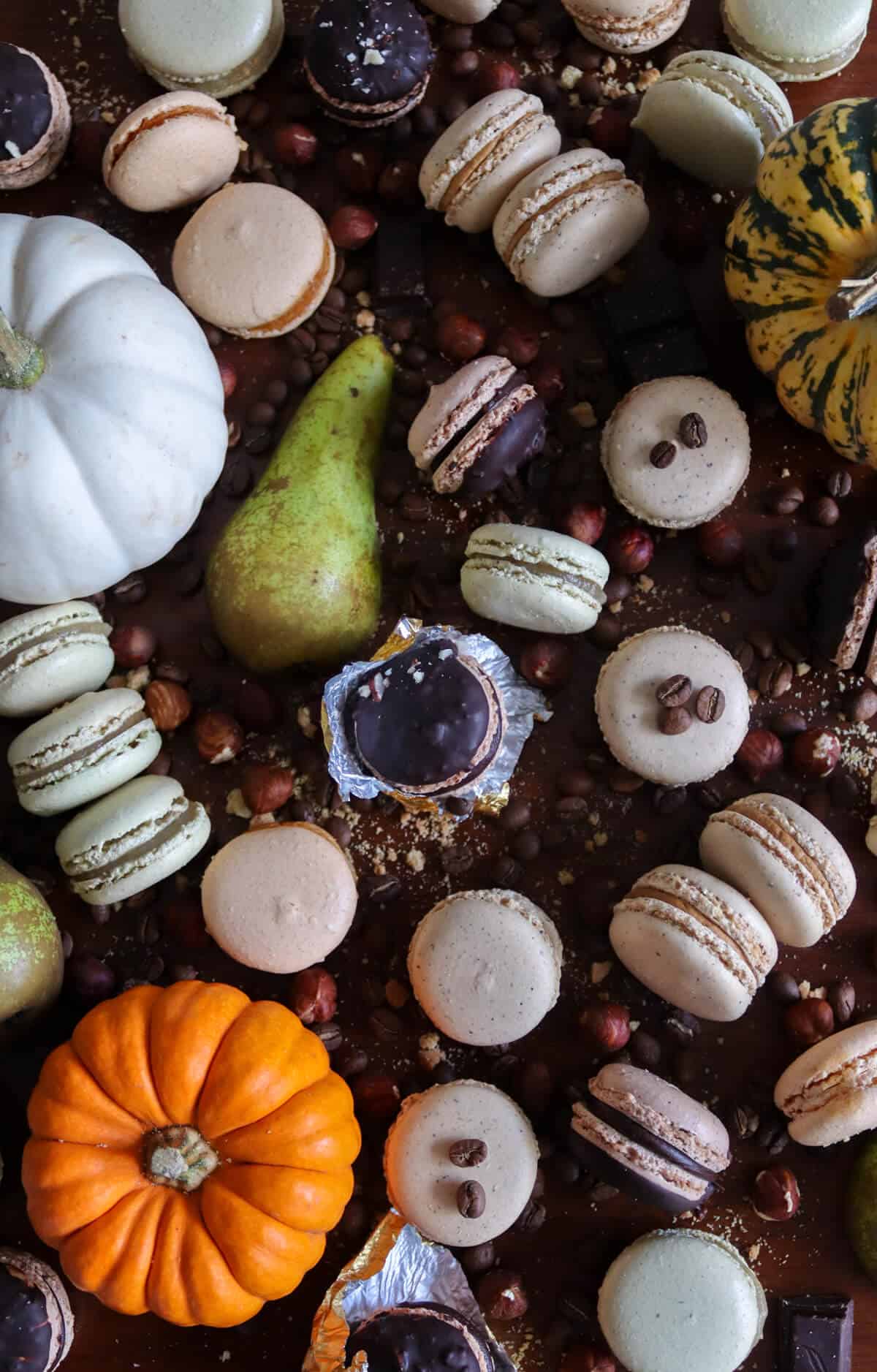Fall macarons displayed on a table surrounded by pumpkins, nuts, pears and coffee beans.
