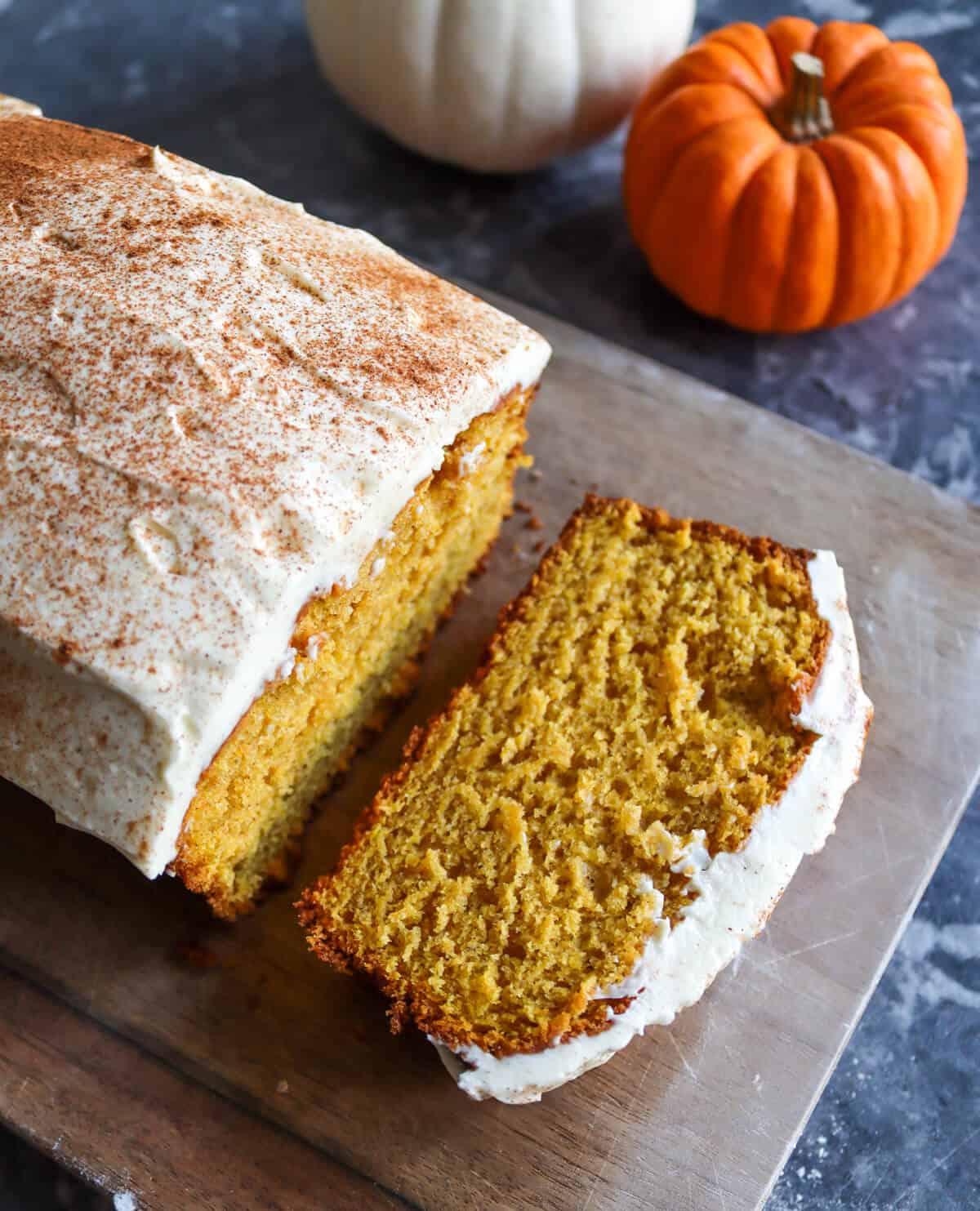 A spiced pumpkin yogurt cake covered in mascarpone cream cheese frosting and dusted with cinnamon, on a bread board surrounded by ornamental pumpkins.
