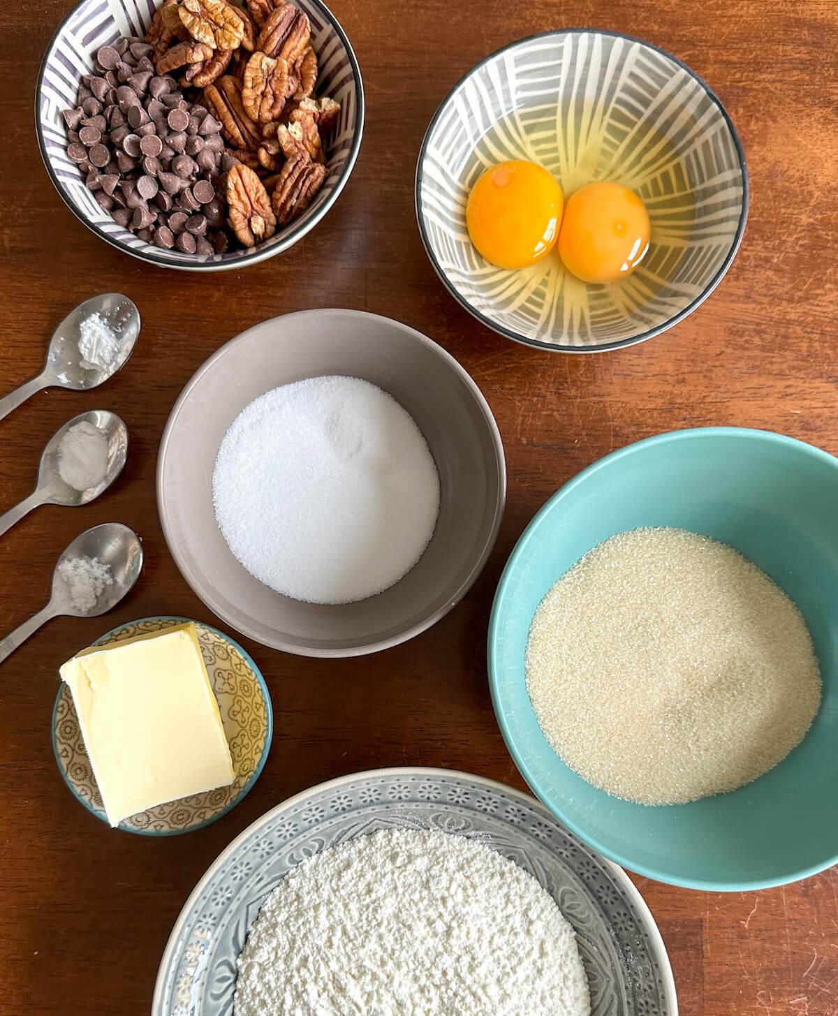The ingredients for brown butter pecan cookies in bowls on a table. Egg yolk and whole egg, brown sugar, white sugar, flour, butter, pecans, chocolate chips, salt, baking powder and baking soda. 