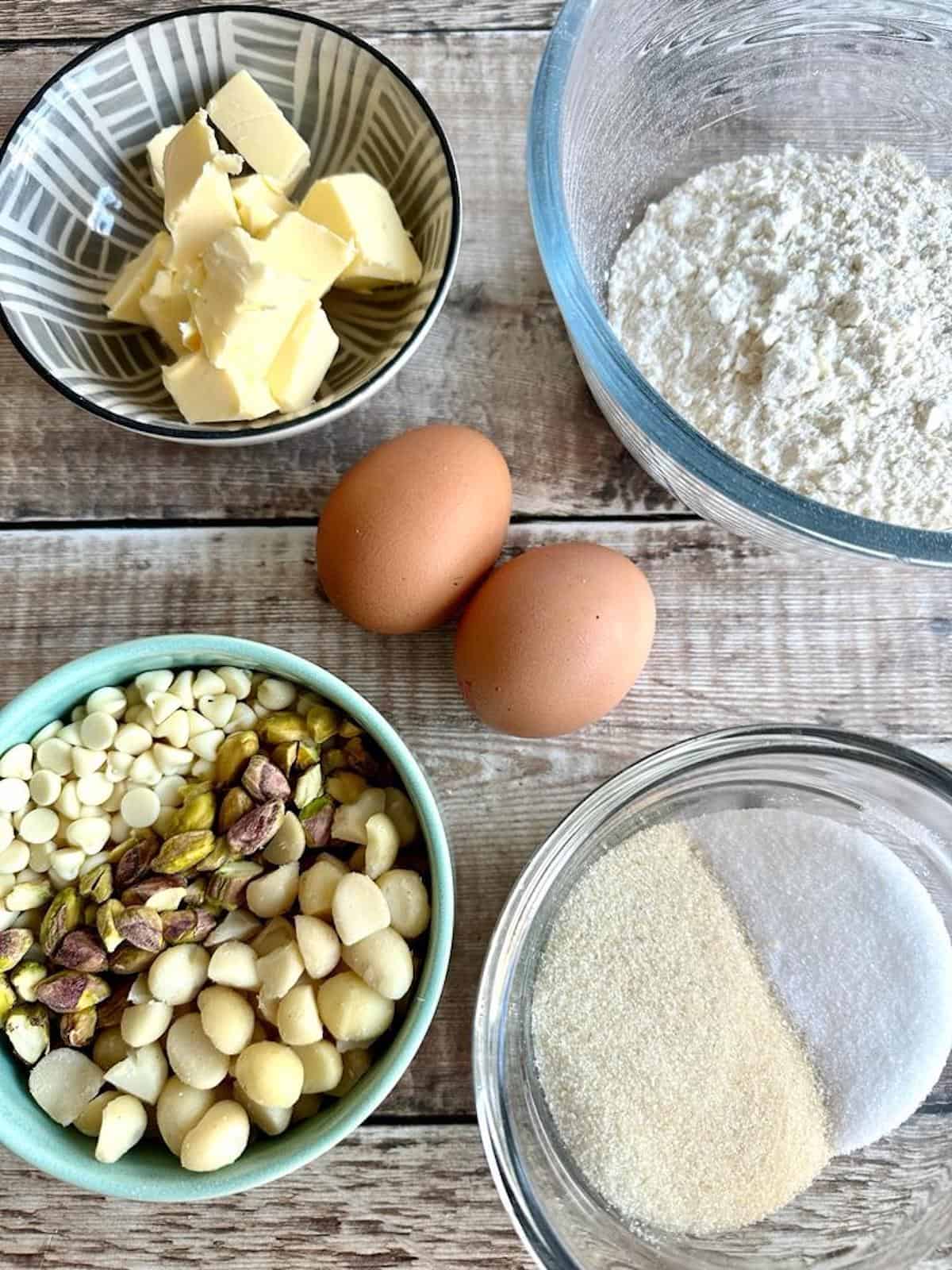 Cookie ingredients in bowls on a table. Butter, flour, eggs, baking powder, baking soda, brown sugar, white sugar, white chocolate chips, pistachio nuts and macadamia nuts. 