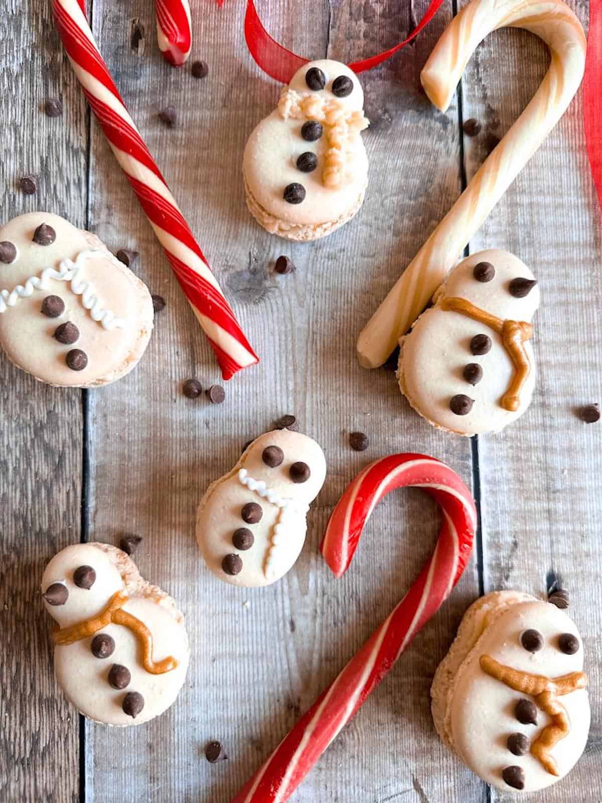 Snowman shaped macrons on a wooden table decorated with chocolate chips and icing, surrounded by candy canes. 