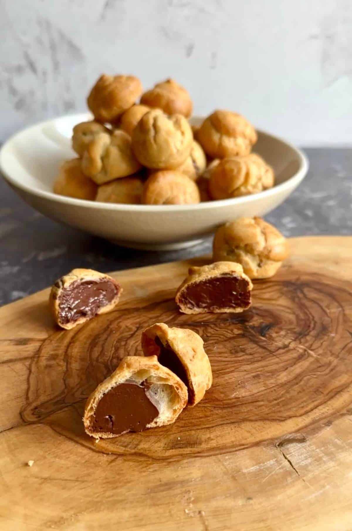 Chocolate classic profiteroles on a wooden chopping board cut in half to show their filling.