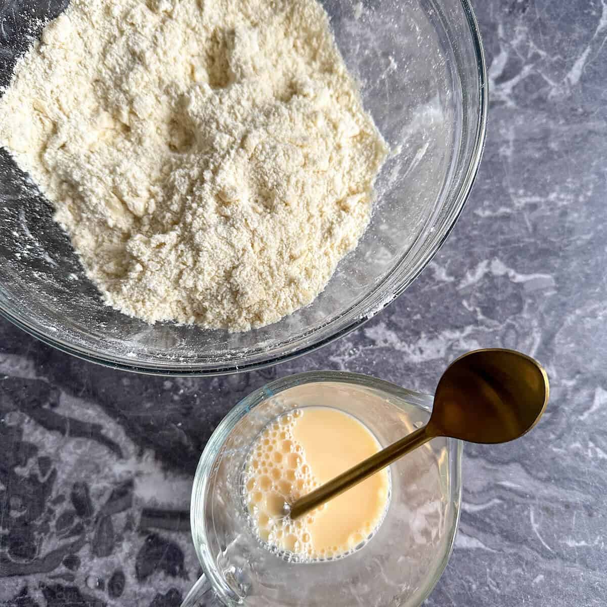 A large mixing bowl with a flour and butter mixture in it that has the consistency of breadcrumbs. Next to the bowl is a small jug with egg yolk and water in it.  