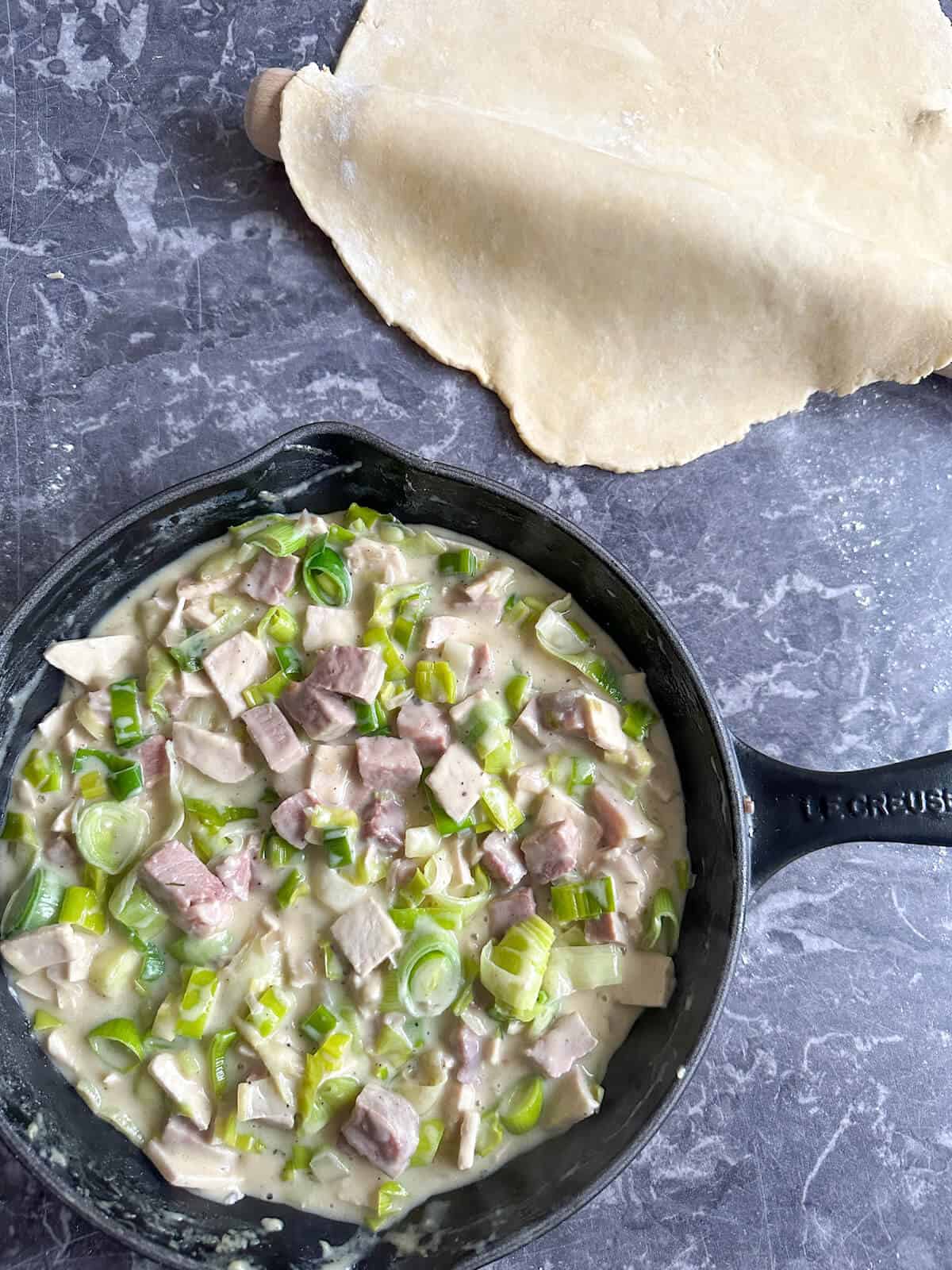 A cast iron skillet with turkey, leek and ham pot pie filling in it, next to a large piece of rolled out pie crust dough.