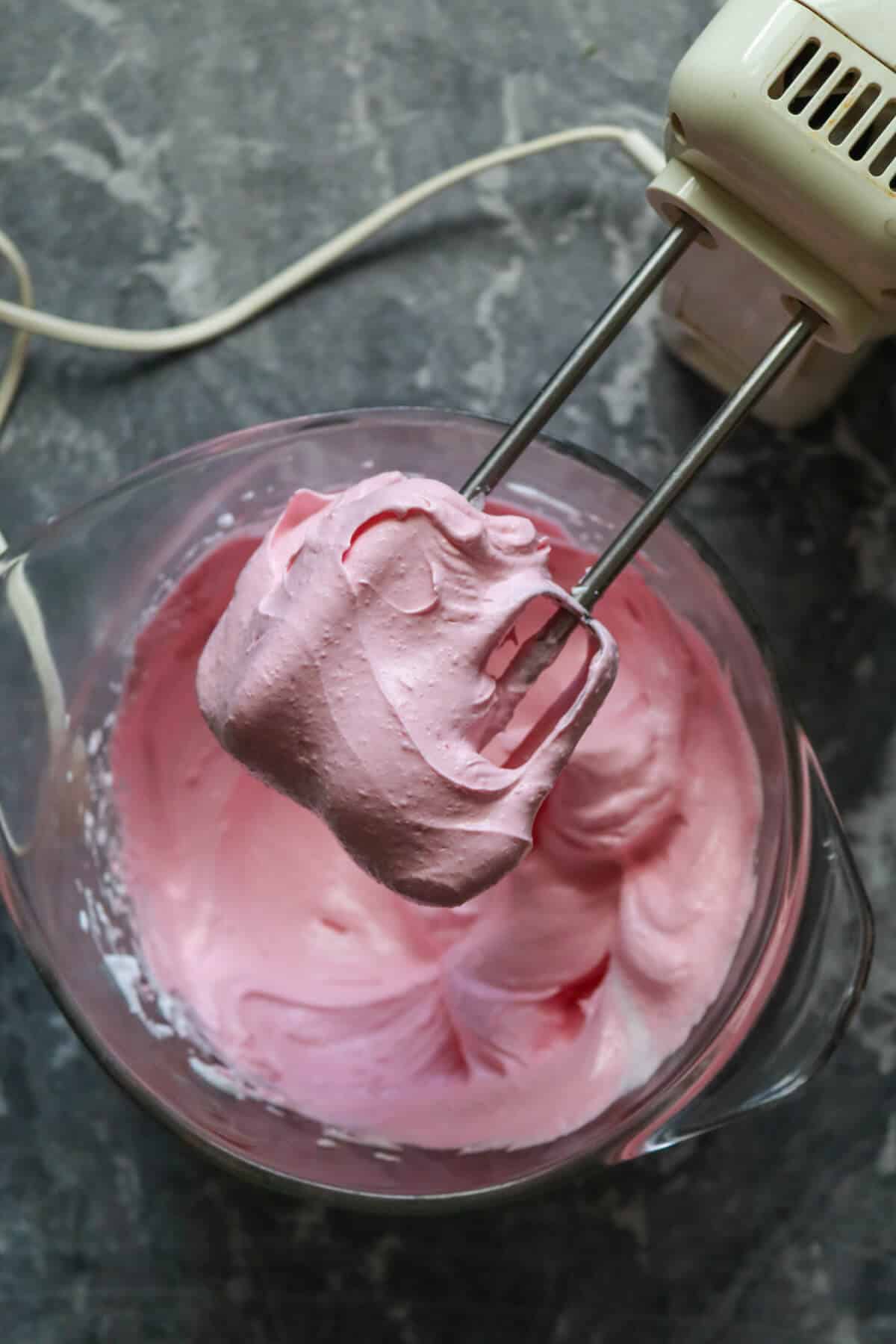 Pink meringue in a mixing bowl with electric hand whisk.