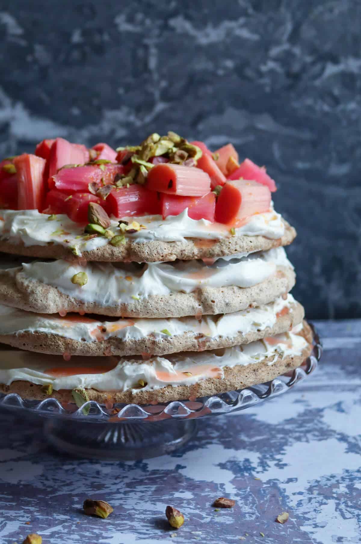 Rhubarb and pistachio pavlova on a cake stand with whole pistachio nuts scattered on top. 