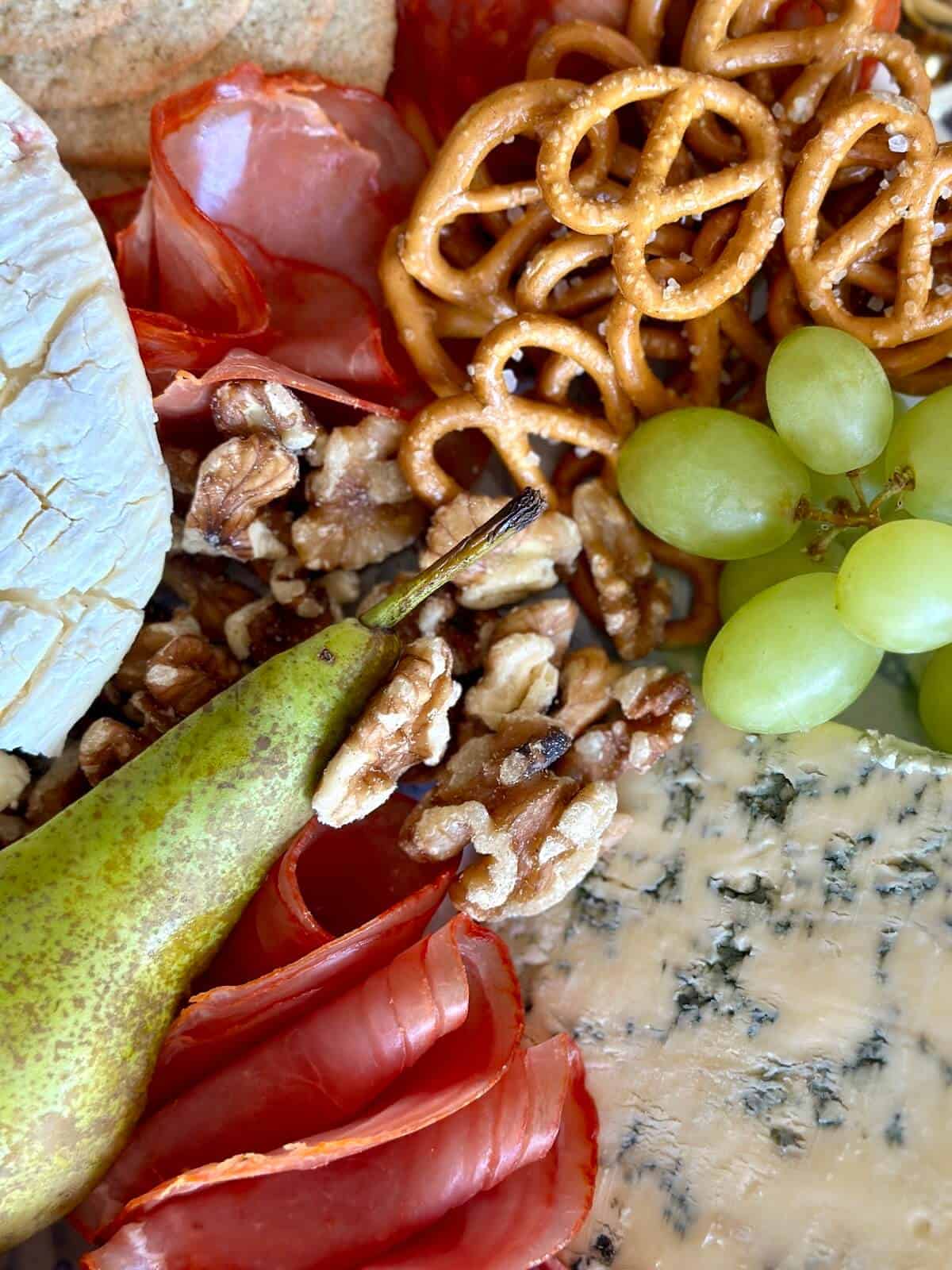 A close up of a cheese and charcuterie board with stilton, pretzels, cold meats, pear and grapes. 