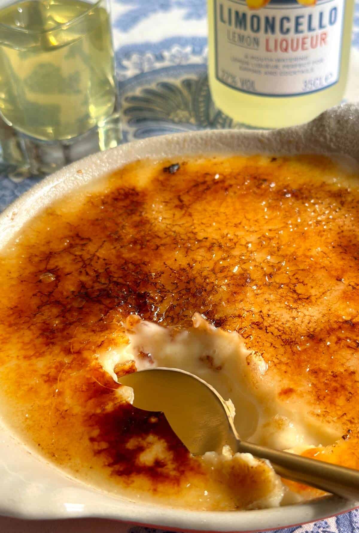 A shallow ramekin of Limoncello creme brulee next to a bottle and glass of Limoncello liqueur. 