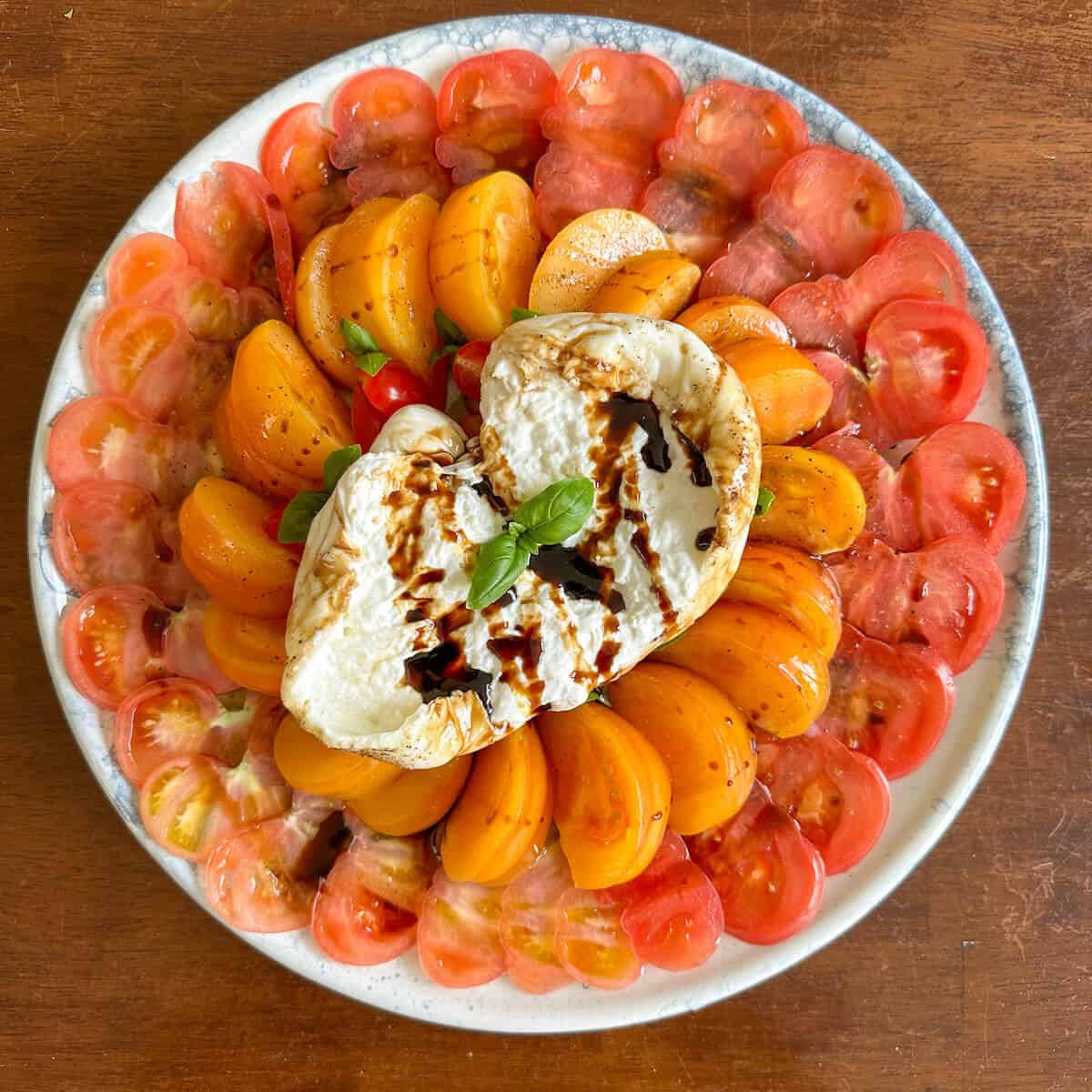 A round plate with sliced heirloom tomatoes and a whole burrata covered in a balsamic glaze and fresh basil. 