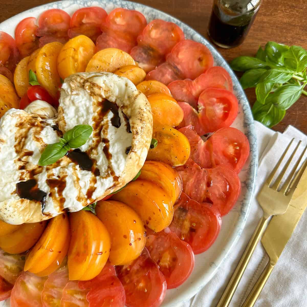 A platter of burrata salad with fresh tomatoes and basil next to a knife snd fork.
