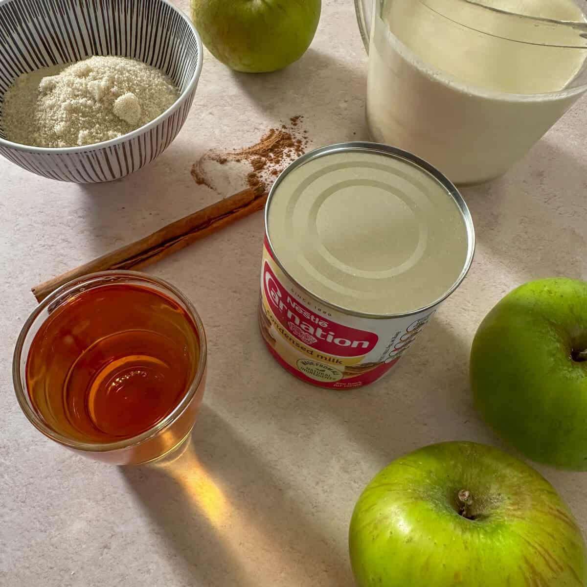 The ingredients for cinnamon bourbon apple ice cream on a table. Brown sugar, cream, bourbon, cinnamon, apples and sweetened condensed milk.