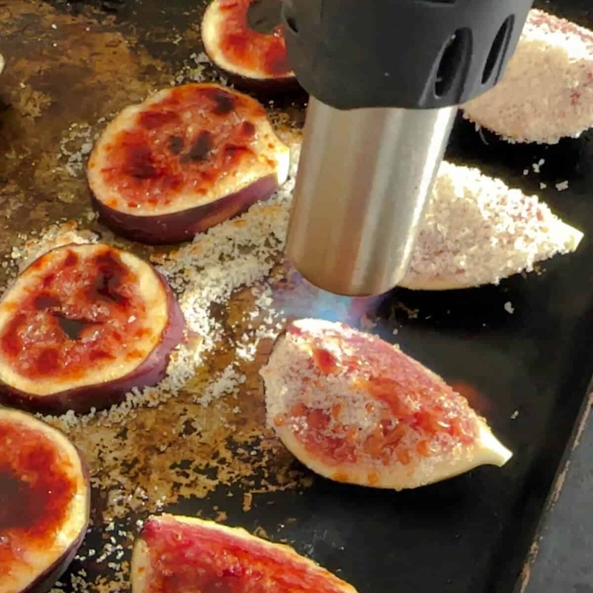 A kitchen blow torch bruleeing figs on a baking tray. 