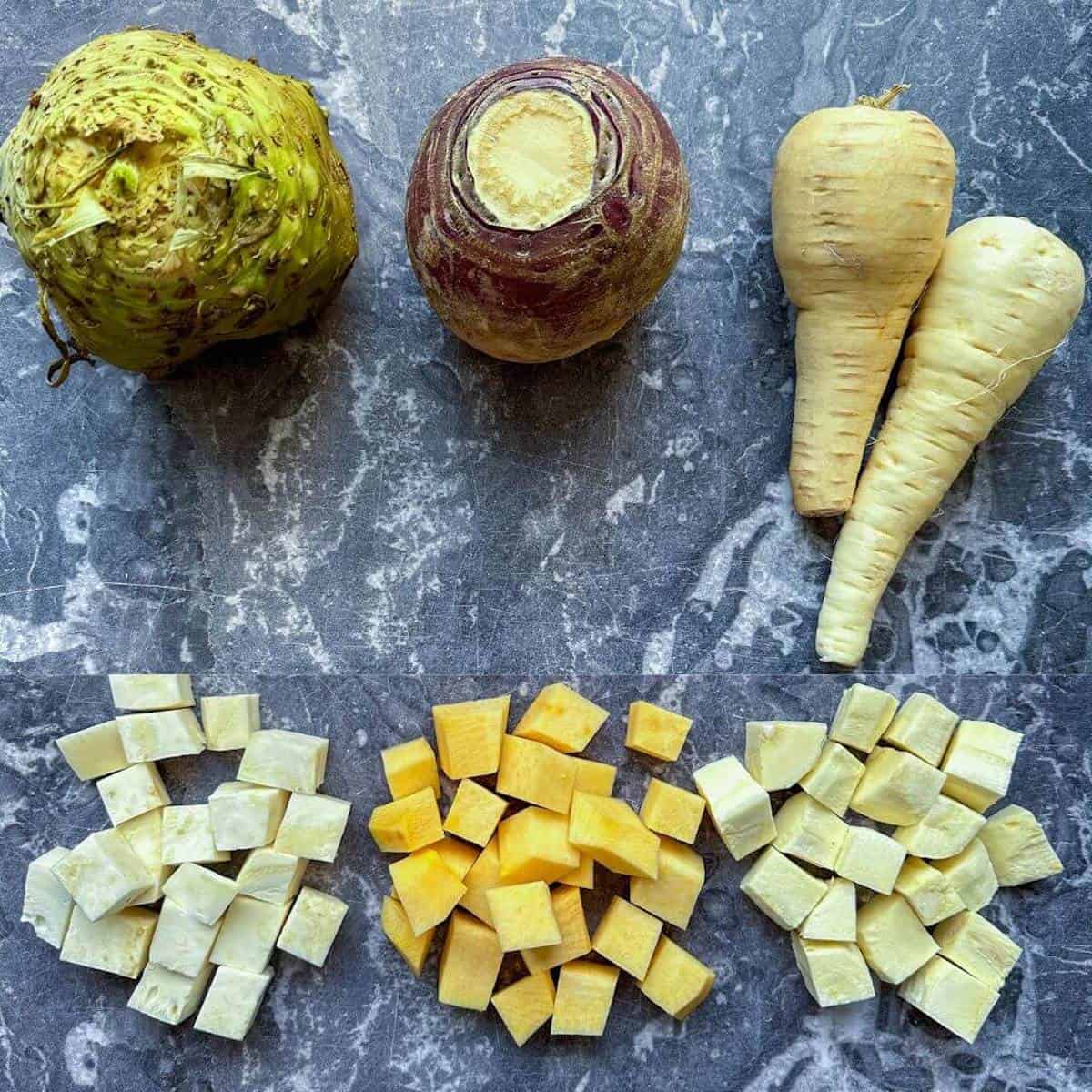 Similar sized cubes of celeriac, swede and parsnip next to the whole unpeeled root vegetables. 
