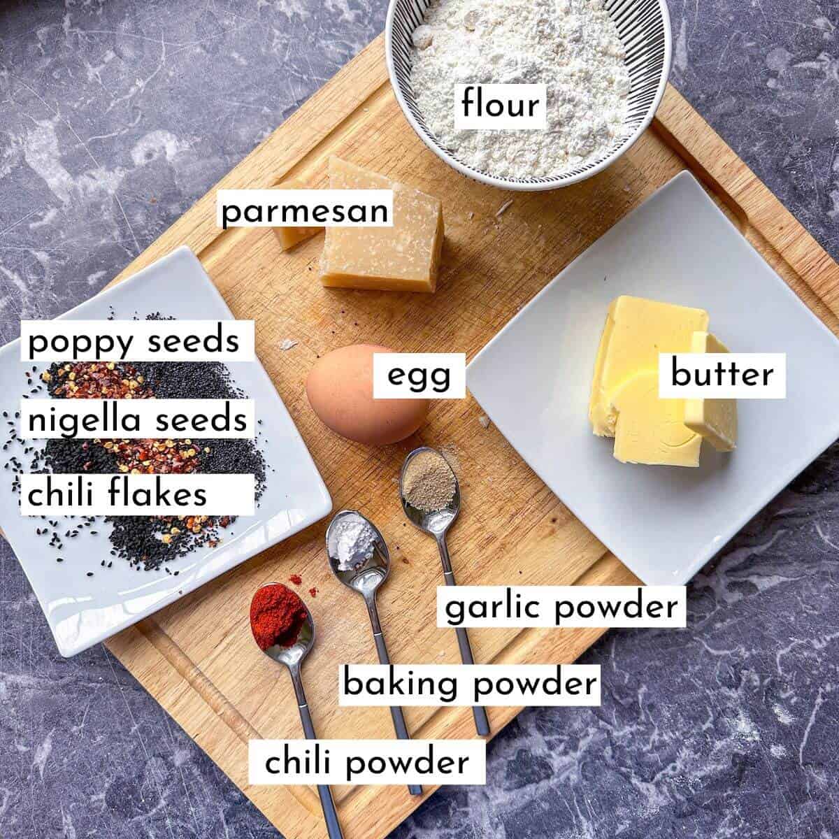 Ingredients for savory parmesan cookies laid out on a worktop. Flour, egg, butter, baking powder, chili powder, garlic powder, parmesan, nigella seeds and chili flakes. 