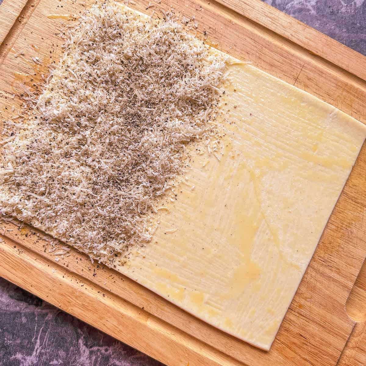 An open piece of puff pastry with grated parmesan cheese and black pepper covering one side of it. 