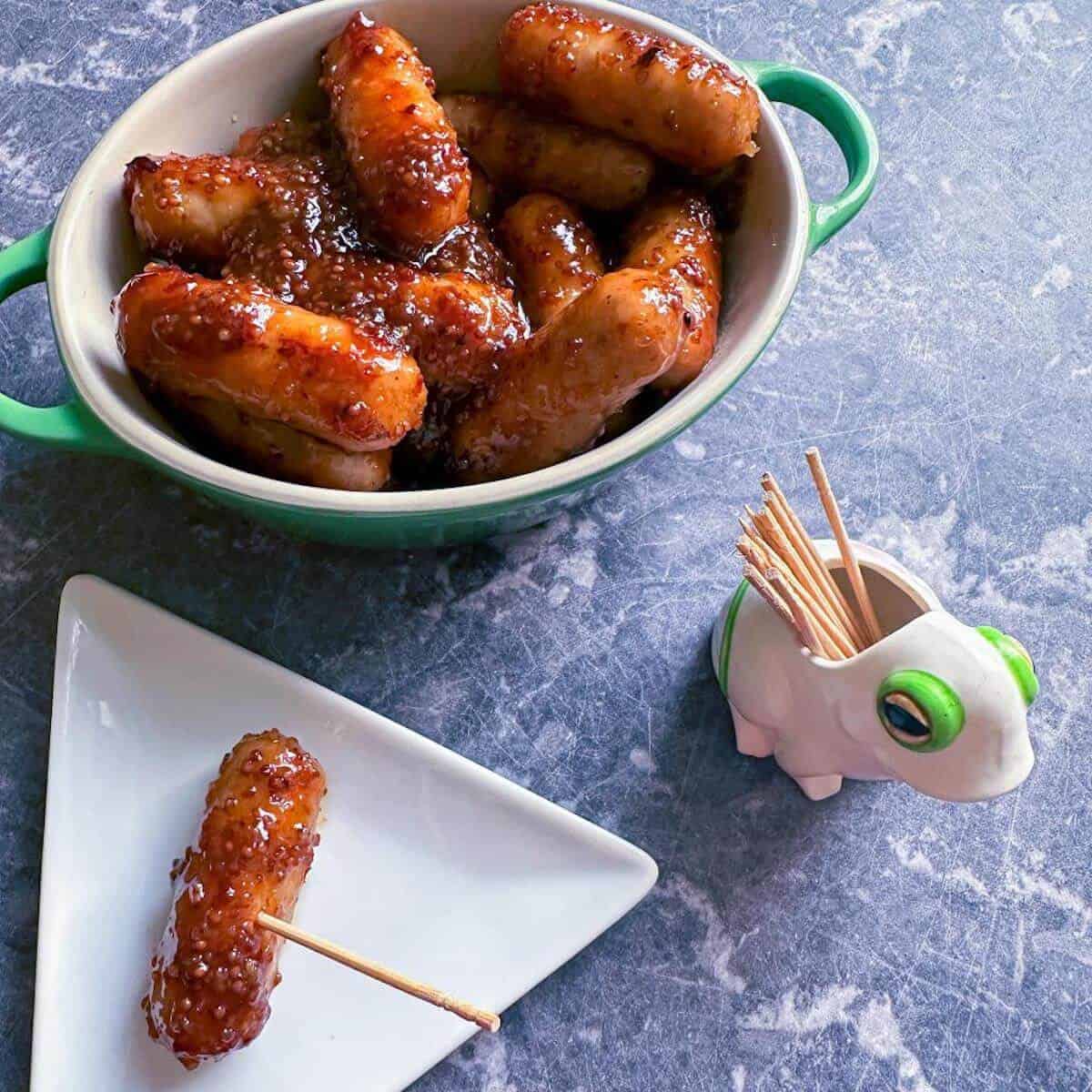 Sticky glazed honey mustard cocktail sausages in a serving bowl next to a pot of cocktail sticks. 