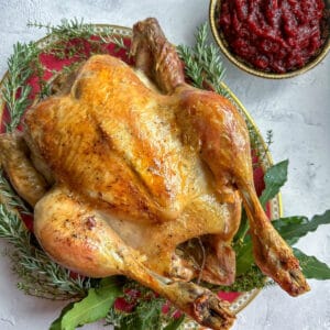 A whole roast capon chicken on a serving platter surrounded by herbs, next to a bowl of cranberry sauce.