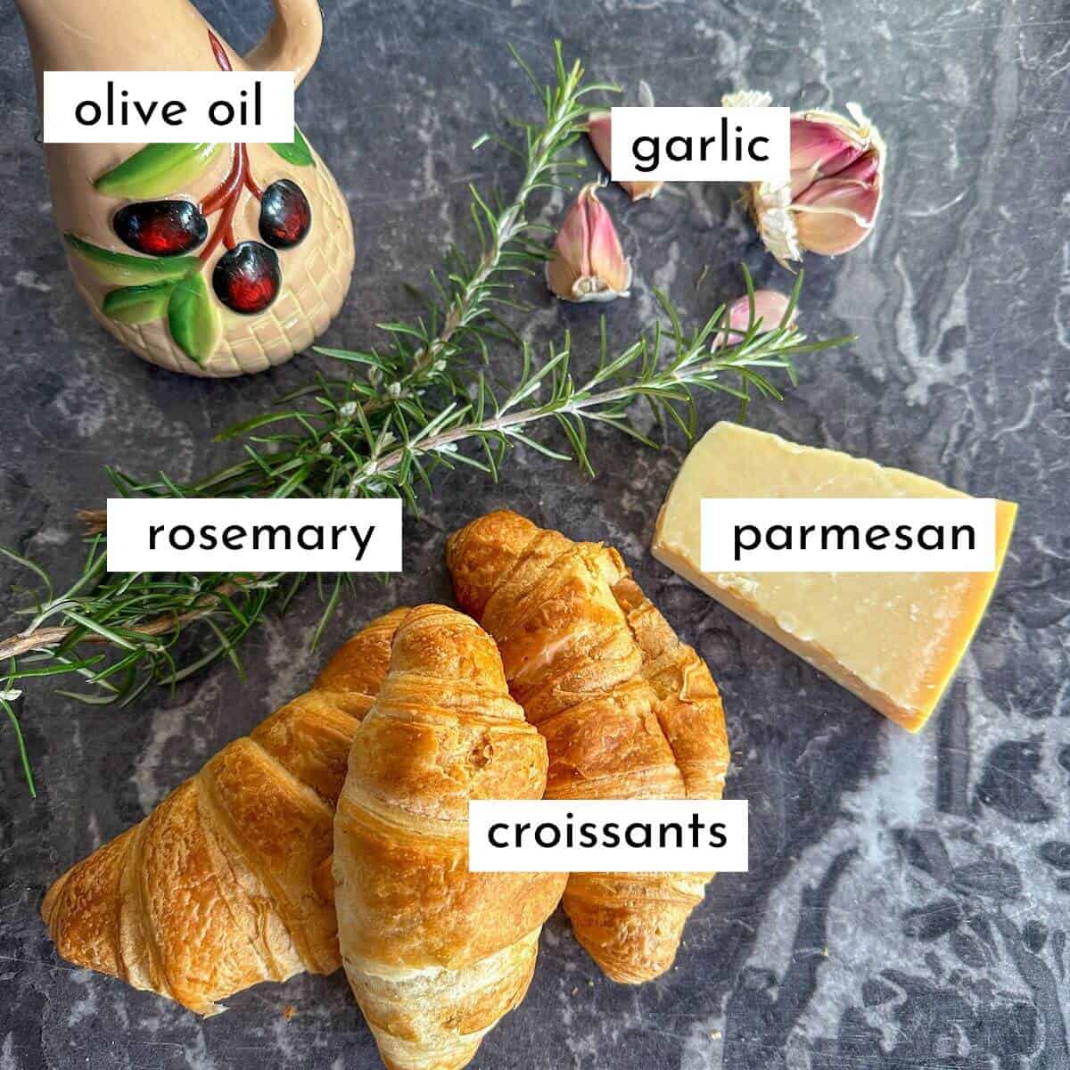 The ingredients for croissant croutons on a worktop. Croissants, parmesan cheese, fresh rosemary, garlic cloves and a jug of olive oil. 