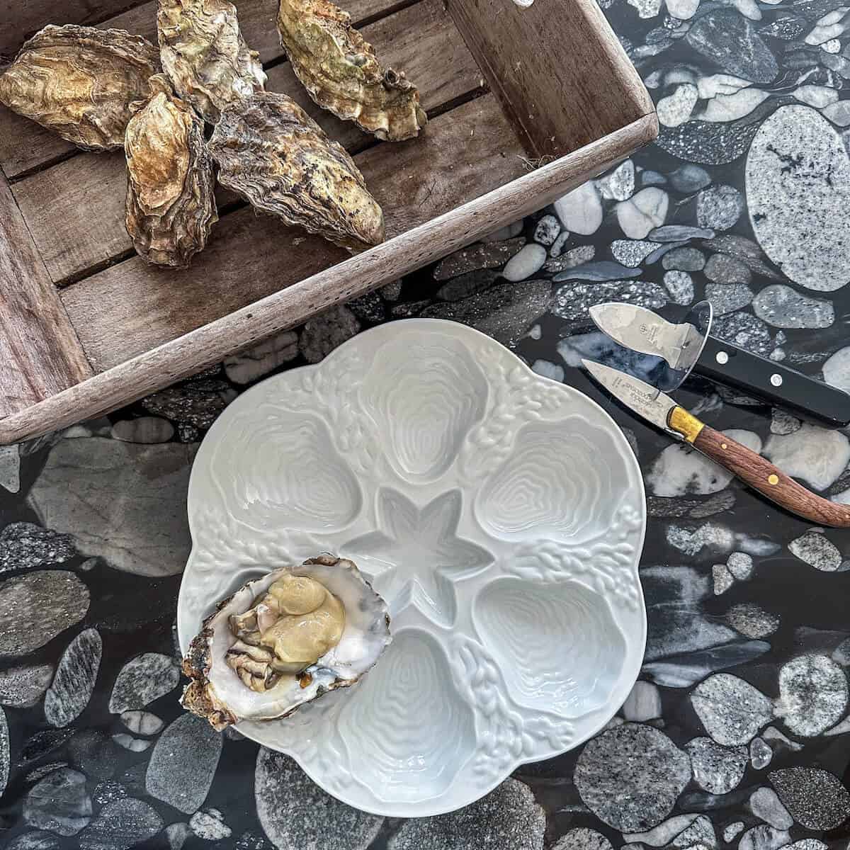 A platter with one shucked oyster on it, next to a crate of fresh oysters. 