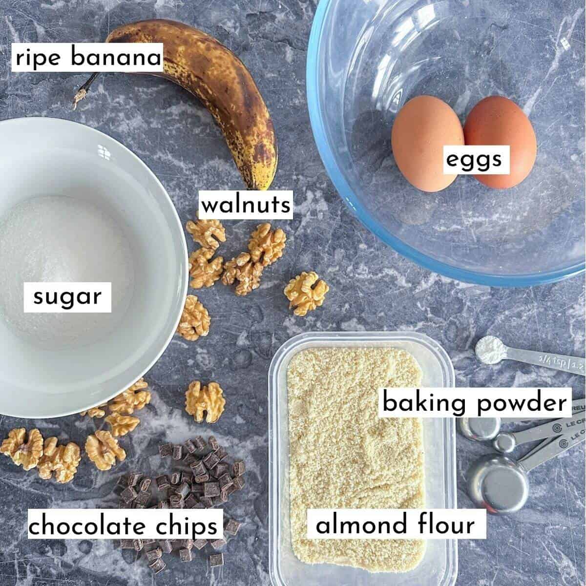 Ingredients for banana sponge pudding on a worktop. Banana, sugar, flour, almond flour, eggs, chocolate chips, walnuts and baking powder. 