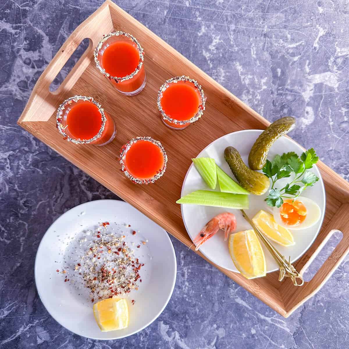 A tray of bloody mary shots next to a plate with garnishes including prawns and boiled egg.