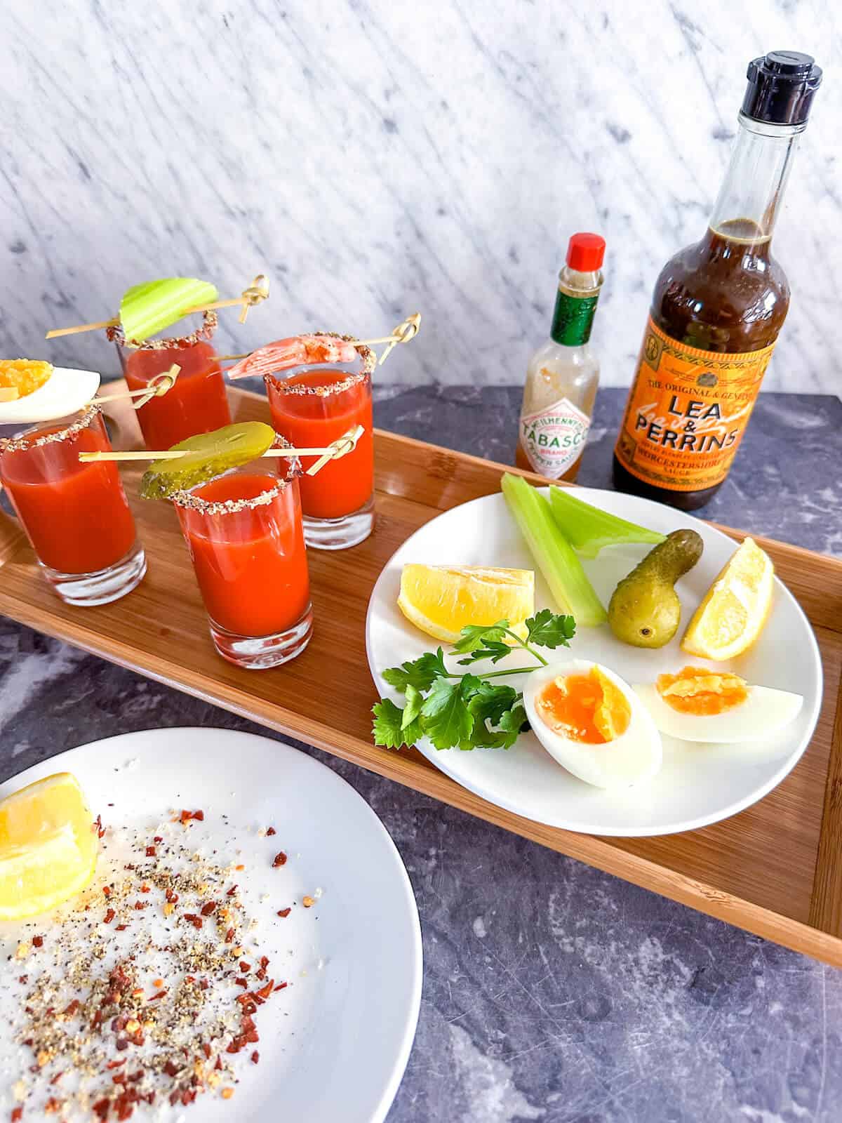 A tray of Bloody Mary amuse bouche shots next to a plate of garnishes, a bottle of Tabasco and a bottle of Worcestershire sauce.