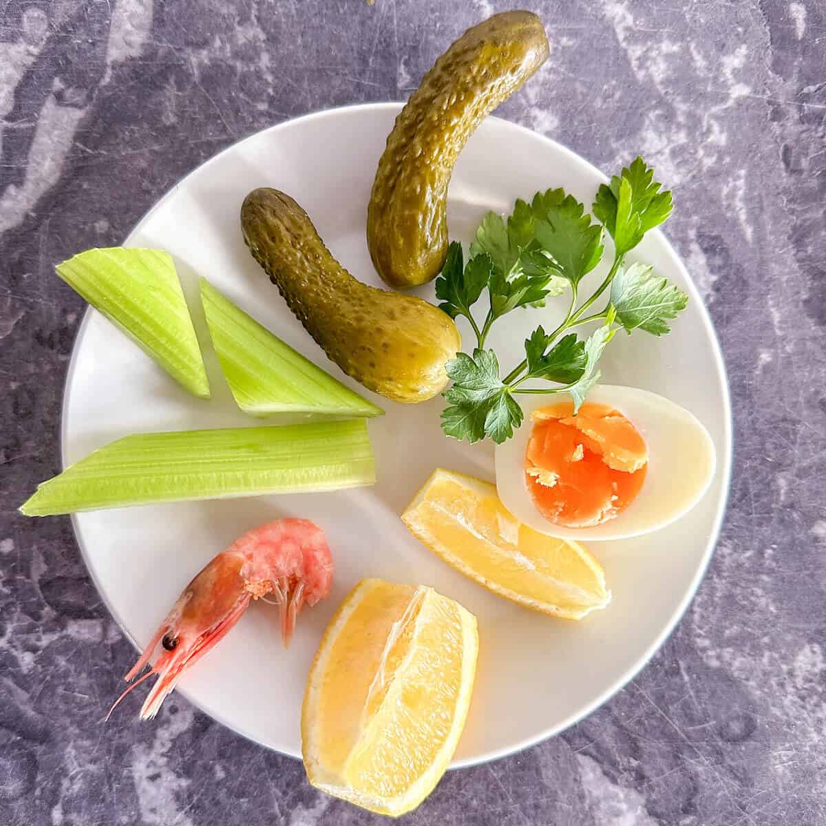 A plate with pickles, prawns, boiled eggs, lemon wedges and celery sticks. 