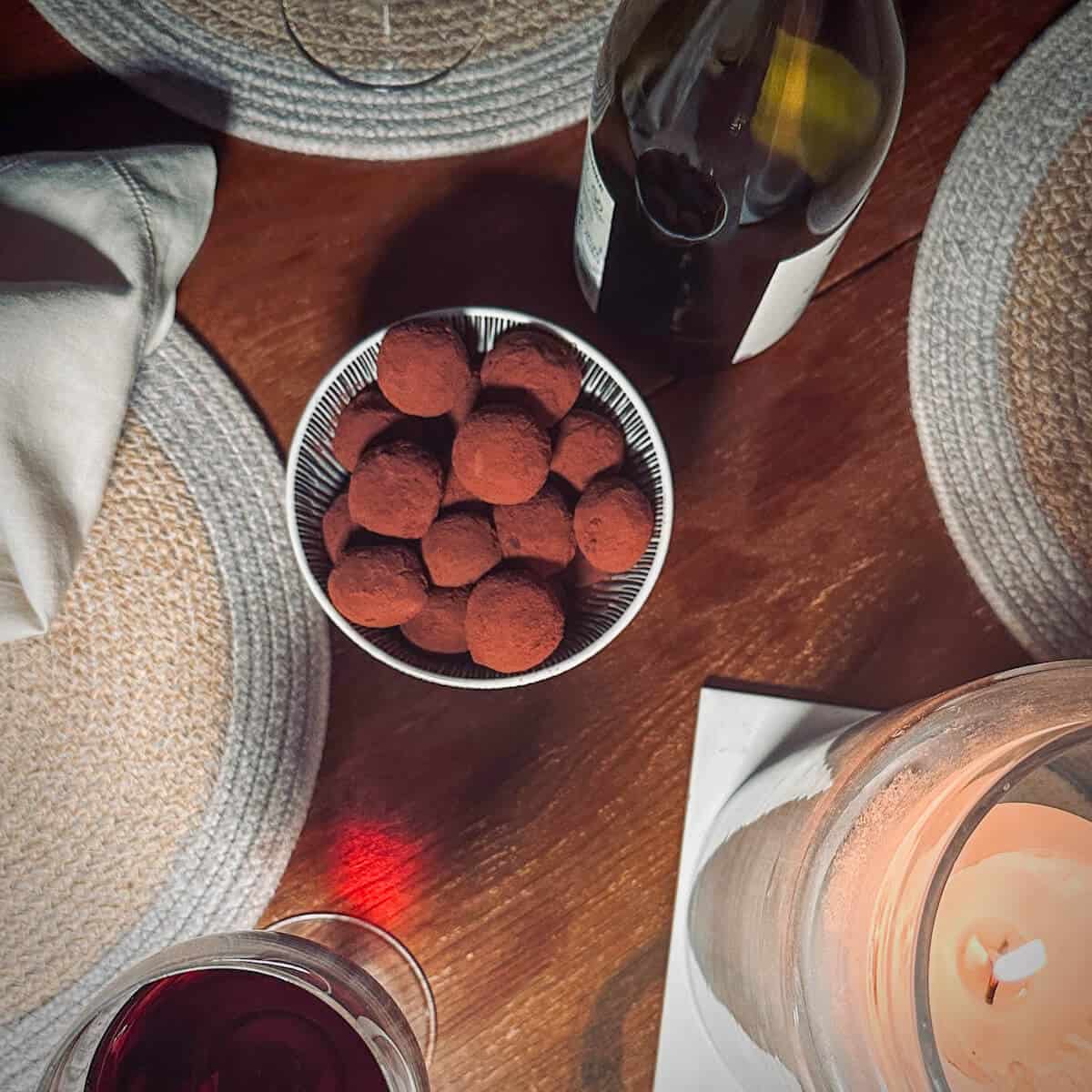 A dining table with a bowl of chocolate truffles, wine glasses, napkins and a bottle of wine. 