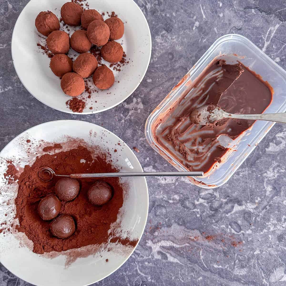 A bowl of cocoa powder with 3 chocolate truffles in it, next to a container of truffle mixture and a plate of coated truffles. 