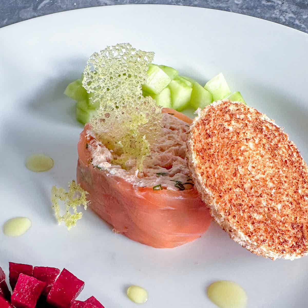 A serving of smoked fish terrine with a coral tuille and slice of toasted bread. 