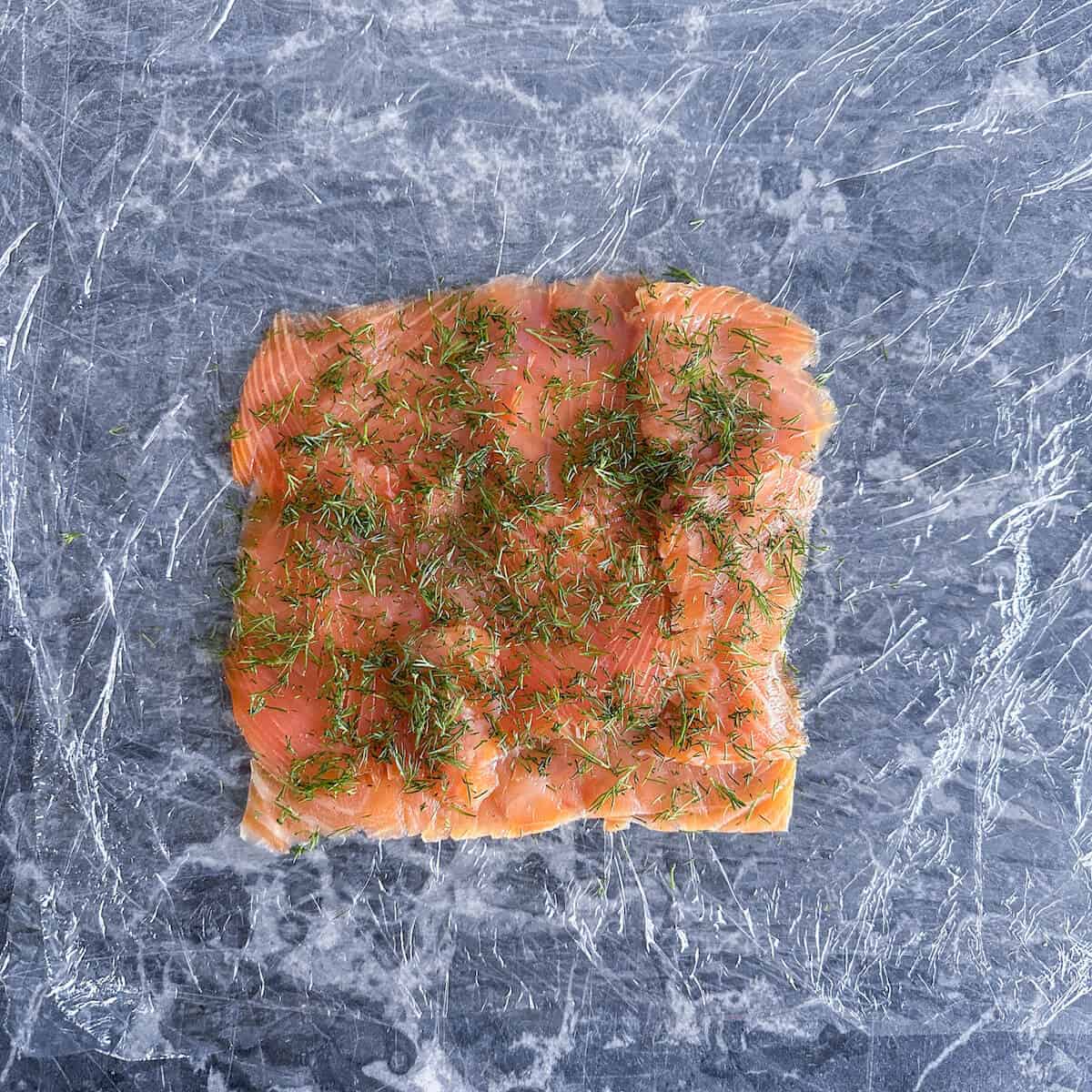 Slices of smoked salmon and fresh dill laid on top of a piece of plastic food wrap. 