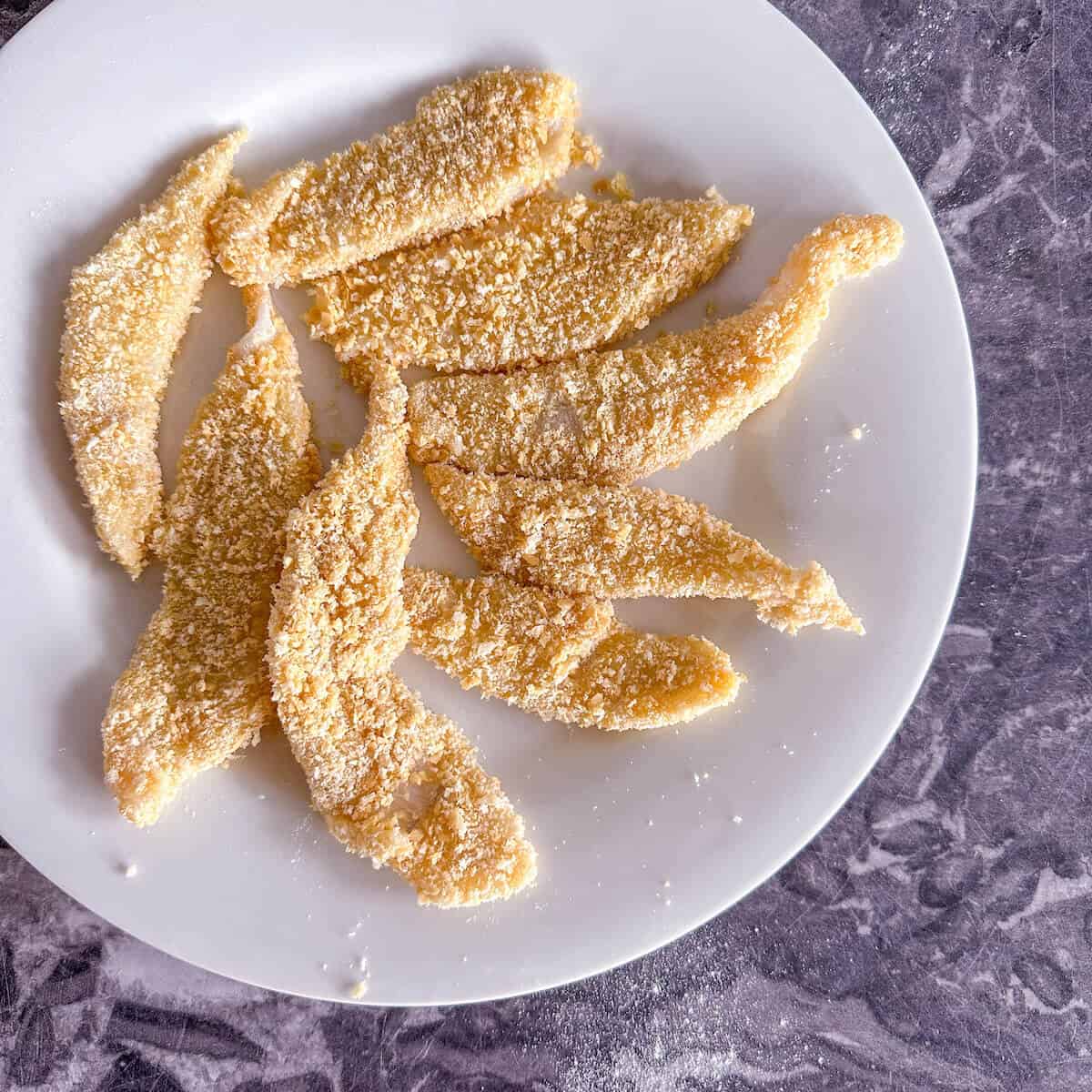 Breadcrumbed raw fish goujons on a plate, ready to be deep fried. 