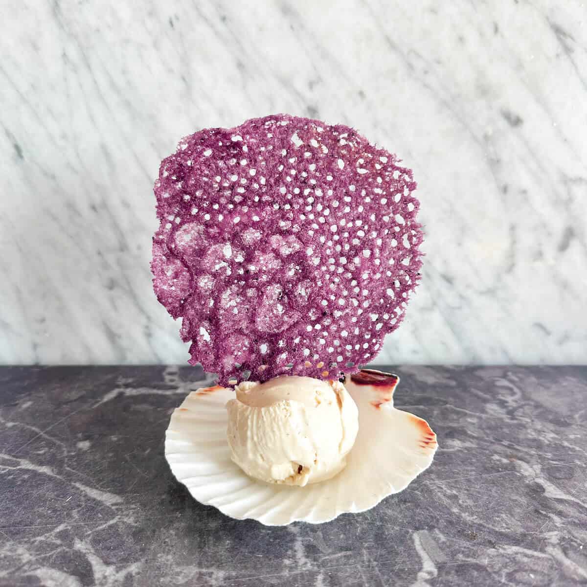 A purple coral tuile garnish on a scoop of ice cream served on a shell.