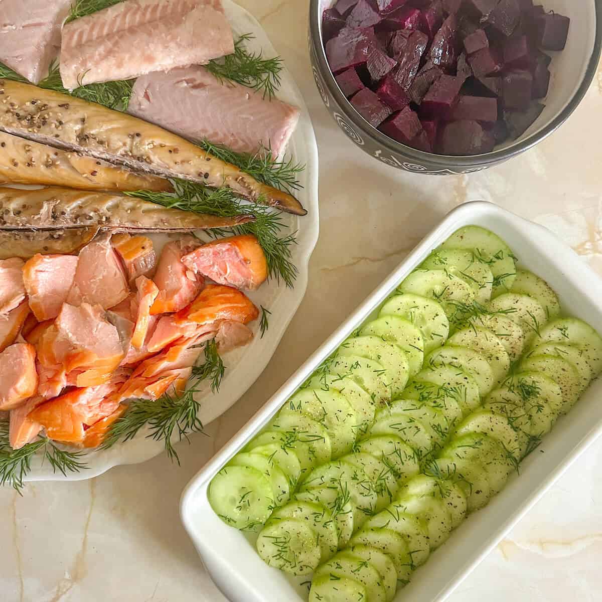 Cucumber salad in a dish next to a bowl of cubed beetroot and a smoked fish platter. 
