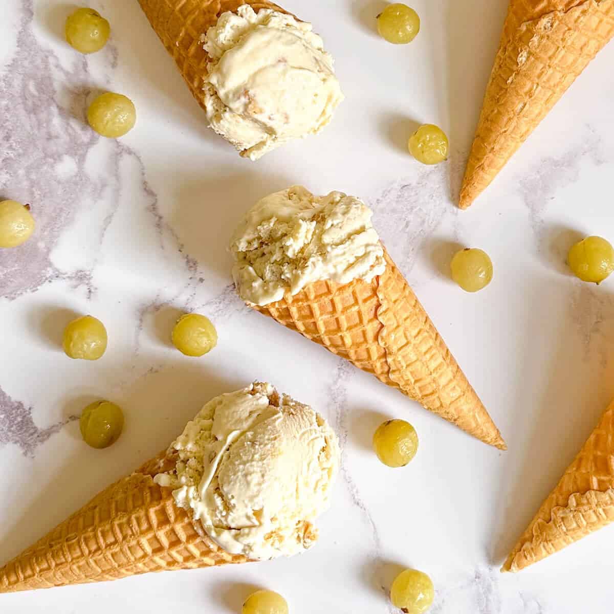 Three ice cream cones of gooseberry ice cream surrounded by gooseberries. 