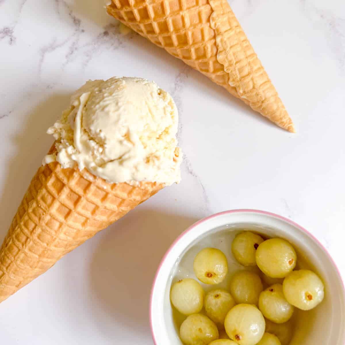 Ice cream cones with a scoop of gooseberry ice cream in, next to a dish of gooseberries. 