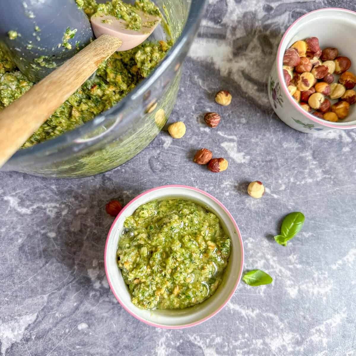 Toasted hazelnut pesto in a ramekin next to a pot of hazelnuts, basil, and a food mixer full of freshly made pesto.